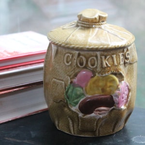 Może przedstawiać: Beżowy ceramiczny słoik na ciasteczka z motywem worka jutowego. Na przodzie napis "COOKIES". Słoik posiada pokrywkę i dekoracyjny otwór odsłaniający kolorowe ciasteczka. W tle stos książek.