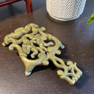 May include: Ornate brass decorative object featuring cherubic figures and scrolling designs. The object has a handle-like extension with a geometric pattern. The brass has a weathered appearance, suggesting age and use. The object is likely a decorative piece.