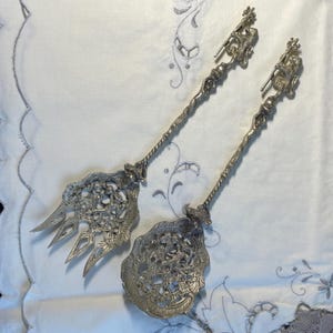 Puede incluir: Dos utensilios de servir plateados ornamentados. Uno es un tenedor con tres púas, el otro una cuchara ranurada. Ambos tienen mangos detallados con una figura en la parte superior y diseños intrincados en los extremos de servir. Sobre un paño blanco bordado.