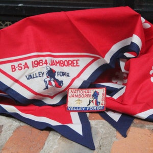Puede incluir: Un pañuelo rojo, blanco y azul de BSA 1964 Jamboree Valley Forge con un parche a juego. El pañuelo tiene un borde blanco con ribete azul y un emblema central. El parche dice "National Jamboree Valley Forge".