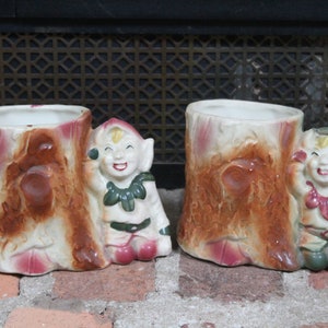 Può includere: Due fioriere vintage in ceramica, ciascuna a forma di ceppo d'albero con una figura di elfo sorridente. Le fioriere hanno un interno bianco, con esterni marroni, rosa e crema. Le figure degli elfi sono bianche, verdi e rosa.