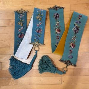 May include: Four teal decorative textile panels with floral and fruit embroidery. Each panel has a gold-coloured metal top and a tassel. Two panels have white backing, and one has a yellow backing.