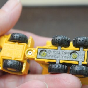 Pode incluir: Um caminh&atilde;o basculante de brinquedo amarelo com rodas pretas. A parte inferior do caminh&atilde;o &eacute; vis&iacute;vel, com o texto "&copy; 2002 TOMY MADE IN CHINA". O caminh&atilde;o &eacute; segurado em uma m&atilde;o.