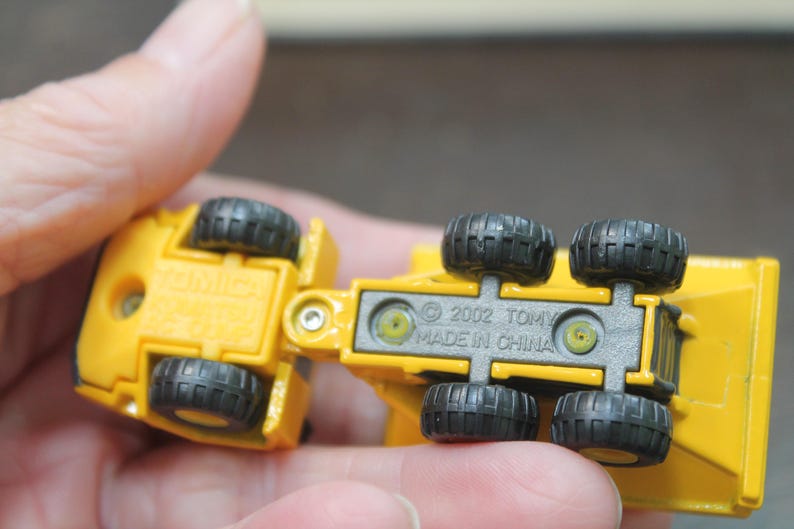 Pode incluir: Um caminh&atilde;o basculante de brinquedo amarelo com rodas pretas. A parte inferior do caminh&atilde;o &eacute; vis&iacute;vel, com o texto "&copy; 2002 TOMY MADE IN CHINA". O caminh&atilde;o &eacute; segurado em uma m&atilde;o.