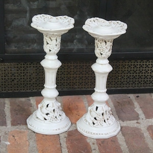 Puede incluir: Dos candelabros blancos y ornamentados con diseños florales y de hojas. Los candelabros tienen una parte superior en forma de corazón y una base detallada. Los candelabros están hechos de un material blanco con detalles dorados.