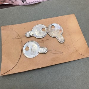 May include: Three vintage metal keys, stamped "MADE IN GERMANY", rest on a brown paper envelope. A pink and white measuring tape is partially visible in the background. The keys have a unique, rounded shape.