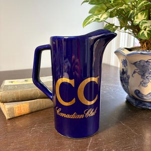 Puede incluir: Jarra azul cobalto con asa y pico vertedor. La jarra presenta las letras doradas "CC" y las palabras "Canadian Club" en escritura dorada. La jarra está sobre una superficie de madera.