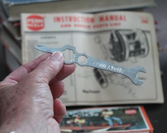 May include: A silver metal wrench tool with the text "Penn Reels" engraved on it. The tool has multiple circular openings and a wrench end. The background includes an instruction manual and a box.