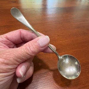 Puede incluir: Una cuchara pequeña de metal plateado con un cuenco redondeado y un mango delgado y liso que se estrecha hacia el extremo. La superficie del metal presenta un acabado pulido con sutiles signos de desgaste y un ligero deslustre.