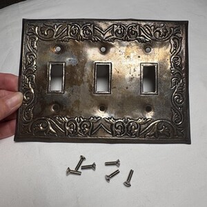 May include: A tarnished silver-colored light switch plate with an ornate floral design around the edges. The plate has three rectangular openings for the switches and several screw holes. Six silver screws are scattered below the plate.
