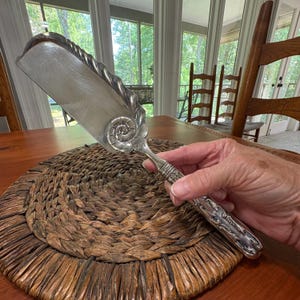 May include: Silver-toned cake server with a decorative handle and a seashell design. The server has a wide, flat blade and a twisted rope design along the edge. The handle features intricate floral and geometric patterns.