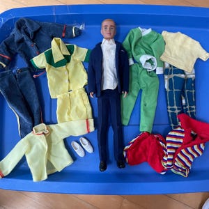 Op de afbeelding: Een pop met diverse outfits in een blauwe plastic bak. Outfits zijn onder meer een marineblauw pak, gele shirts en shorts, een groene broek en shirt, en gestreepte en effen tops. Witte en zwarte schoenen zijn ook inbegrepen.