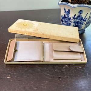 May include: A tan leather wallet with a snap closure, open inside a matching box. The box lid is tan with the words "LADY BUXTON" and a gold sunburst design. The wallet is on a dark brown surface.