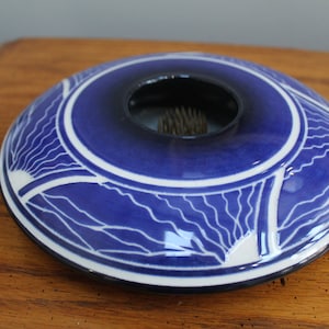 Puede incluir: Un ranero de cerámica azul con una abertura central y un diseño geométrico blanco y azul. El ranero es redondo y tiene un centro azul oscuro. El diseño incluye líneas y formas blancas.