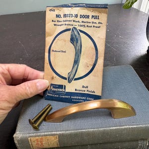 Könnte beinhalten: Ein bronzefarbener Türgriff mit matter Oberfläche, zusammen mit seiner Originalverpackung. Die Verpackung ist beige mit blauem Text und einer Abbildung des Türgriffs. Zwei Messingschrauben sind ebenfalls vorhanden.