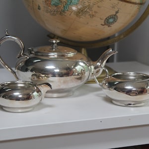 May include: A silver tea set, including a teapot with a lid, a creamer, and a sugar bowl. The set is highly reflective, with curved handles and spouts. The tea set is displayed on a white surface.
