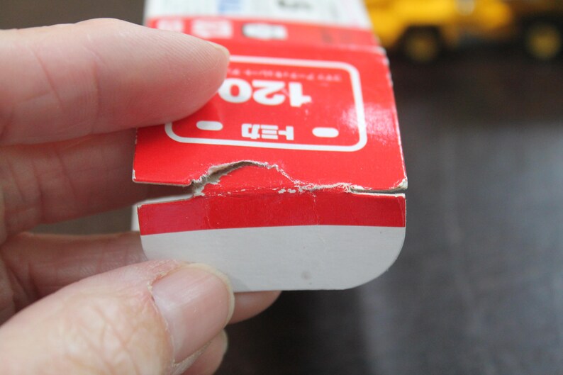 Pode incluir: Uma caixa de brinquedos vermelha e branca com o n&uacute;mero 120 em branco. A caixa tem texto japon&ecirc;s e &eacute; segurada por uma m&atilde;o. A caixa est&aacute; ligeiramente danificada.