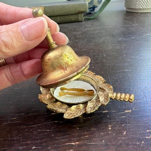以下が含まれることがあります： 小さなハンドルと葉の形をした装飾的なベースが付いた、ヴィンテージの真鍮のベル。ベルの中には、白い表面に置かれた小さな金色のピンセットがあります。ベルは開いた状態で、その内容が明らかになっています。