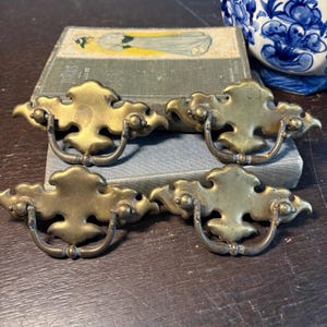 Może przedstawiać: Cztery antyczne mosiężne uchwyty do szuflad o ozdobnym wzorze. Każdy uchwyt ma zakrzywiony uchwyt i stylizowaną, liściastą płytkę tylną. Uchwyty są ułożone na zabytkowej książce, co sugeruje klasyczną estetykę.