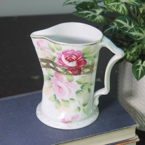 May include: White porcelain creamer with a handle, adorned with pink roses and green leaves. The creamer features a gold band and trim, adding a touch of elegance. A decorative piece for serving cream or displaying.