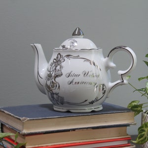 May include: White ceramic teapot with silver accents and the text "Silver Wedding Anniversary." The teapot has a curved spout, handle, and lid. It sits on a stack of books with a plant in the background.