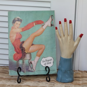 Könnte beinhalten: Ein Vintage-Magazin-Cover mit dem Titel "Ice Vogues of 1954" mit einer Frau in einem roten Kleid und Schlittschuhen. Daneben eine Schaufensterpuppenhand mit rot lackierten Nägeln und einem blauen Ärmel. Das Magazin wird auf einem schwarzen Metallständer präsentiert.