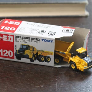 Pode incluir: Um brinquedo de caminh&atilde;o basculante articulado Komatsu amarelo, escala 1/144, est&aacute; parcialmente dentro de sua caixa vermelha e branca. A caixa apresenta a marca "Tomica" e o n&uacute;mero "120". O brinquedo &eacute; uma r&eacute;plica em miniatura.