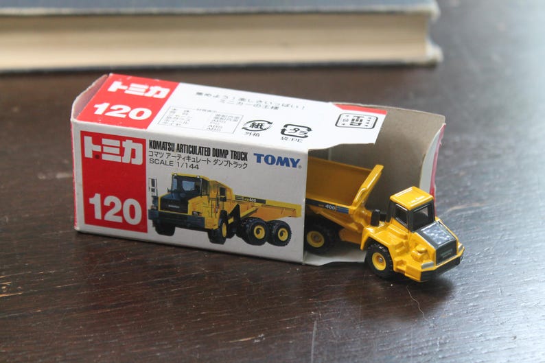 Pode incluir: Um brinquedo de caminh&atilde;o basculante articulado Komatsu amarelo, escala 1/144, est&aacute; parcialmente dentro de sua caixa vermelha e branca. A caixa apresenta a marca "Tomica" e o n&uacute;mero "120". O brinquedo &eacute; uma r&eacute;plica em miniatura.