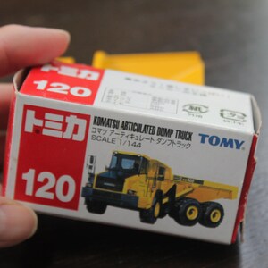 Pode incluir: Um pequeno brinquedo Komatsu Articulated Dump Truck, escala 1/144, em caixa. A caixa vermelha e branca exibe o n&uacute;mero 120. O caminh&atilde;o de brinquedo &eacute; amarelo e preto.