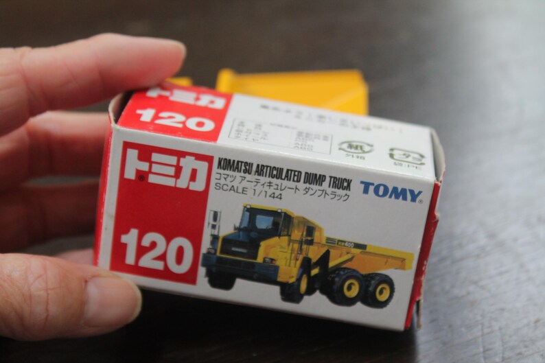 Pode incluir: Um pequeno brinquedo Komatsu Articulated Dump Truck, escala 1/144, em caixa. A caixa vermelha e branca exibe o n&uacute;mero 120. O caminh&atilde;o de brinquedo &eacute; amarelo e preto.