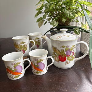 Pode incluir: Um conjunto de chá de cerâmica branca com uma chaleira e quatro canecas, cada uma adornada com ilustrações coloridas de vegetais. A chaleira tem uma tampa com desenhos de cogumelos. As canecas e a chaleira têm uma borda verde.