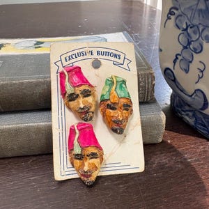 Op de afbeelding: Drie vintage gezichtsvormige knopen met rode en groene hoeden worden getoond op een kaart met de tekst "EXCLUSIVE BUTTONS". De knopen hebben gedetailleerde gelaatstrekken en zijn waarschijnlijk gemaakt van een geschilderd materiaal.