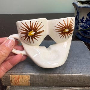 May include: A white ceramic double-cup vessel featuring two conjoined bowls with a single handle. Each bowl is decorated with a brown starburst pattern with a yellow center. The piece has a smooth, glossy finish and a unique, asymmetrical shape.
