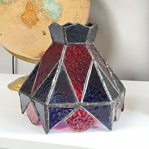 May include: A stained glass lampshade with a geometric design. The shade features red, blue, and purple glass panels, creating a colourful and decorative accent. The lampshade is designed with a unique shape and is suitable for a small lamp.