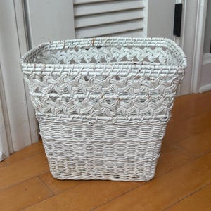 May include: A white square woven basket with a decorative pattern around the top. The basket is made of a natural material and has a sturdy, open design. The basket is suitable for storage.