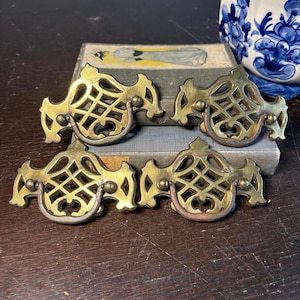 May include: Four antique brass drawer pulls with an ornate, openwork design. Each pull features a curved handle and decorative detailing. The pulls are arranged on a wooden surface, suggesting a vintage or antique theme.