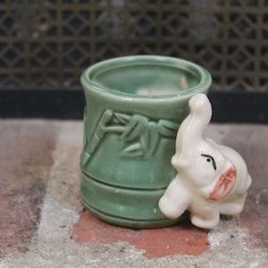 May include: A small, green ceramic container with a bamboo design and a small, off-white elephant figurine attached. The elephant has pink accents on its ears and trunk. The container is sitting on a brick surface.