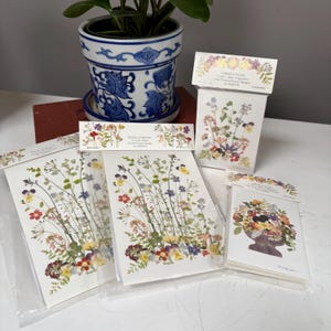 May include: A collection of stationery items featuring pressed flower designs. The items include score pads, bridge tallies, and notecards. The designs showcase a variety of colorful flowers and foliage. A blue and white floral pot with a plant is in the background.