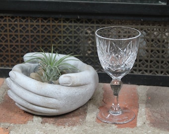 May include: A cut crystal wine glass stands on a brick surface. Next to it, a grey stone planter shaped like cupped hands holds a small plant and pebbles. The background features a decorative metal grate. The glass is approximately 15 cm tall.
