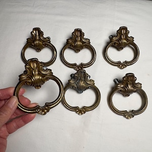 May include: Six antique brass drawer pulls with ornate, scrollwork designs. Each pull features a decorative backplate and a circular handle. The pulls are a warm, aged brass colour, suggesting vintage or retro styling. Ideal for furniture restoration or adding a classic touch.