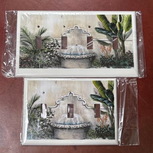 May include: Two decorative light switch covers featuring a detailed illustration of a fountain with lush greenery. The design includes a stone fountain with water cascading into a basin, surrounded by tropical plants and palm trees. The covers have three switch openings.