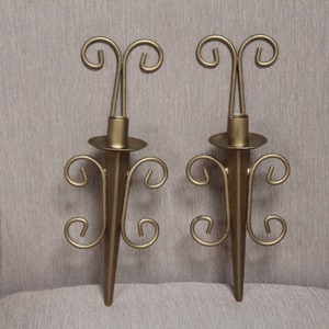 May include: Two gold-colored metal wall sconces. Each sconce has a central spike with a circular candle holder and decorative scrollwork. The sconces are symmetrical and designed to hold candles, adding a touch of elegance to any space.