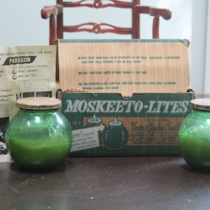 Op de afbeelding: Twee groene glazen kaarsenhouders met kurken deksels en een doos "Moskeeto-Lites" kaarsen. Een vintage advertentie voor "Magic Paragum Base" kaarsenhouders is ook zichtbaar. De items zijn gerangschikt op een donker oppervlak.
