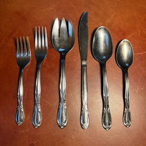 May include: A collection of six silver-coloured cutlery items: two forks, a salad fork, a knife, and two spoons. The handles feature a floral pattern. The cutlery is arranged on a brown surface.