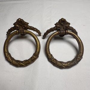 Può includere: Due maniglie per cassetti antiche color bronzo. Ogni maniglia presenta un'impugnatura circolare con un motivo a foglie decorative e una parte superiore ornata e stilizzata. Le maniglie sono progettate per mobili e hanno un'estetica vintage.