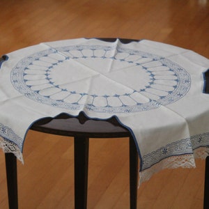May include: A white round tablecloth with a blue floral design and scalloped edges. The tablecloth is placed on a dark brown table. The edges of the tablecloth have a blue trim and lace.