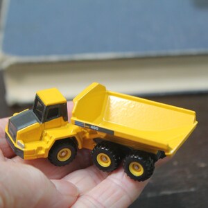 Pode incluir: Um cami&atilde;o basculante de brinquedo amarelo e preto. O cami&atilde;o tem uma cabine amarela e uma caixa basculante amarela. As rodas s&atilde;o pretas. O modelo &eacute; segurado em uma m&atilde;o.