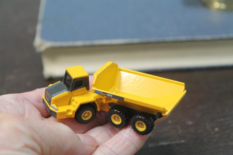 Pode incluir: Um cami&atilde;o basculante de brinquedo amarelo e preto. O cami&atilde;o tem uma cabine amarela e uma caixa basculante amarela. As rodas s&atilde;o pretas. O modelo &eacute; segurado em uma m&atilde;o.