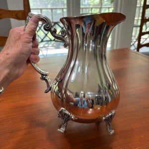 May include: A polished silver-plated pitcher featuring a reflective, curved body with vertical fluting. The vessel has an ornate, scrolled handle and stands on three decorative, claw-style feet. The surface is highly reflective, showing surroundings.