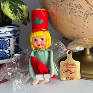 Può includere: Una bambola natalizia vintage con capelli gialli, occhi blu e un cappello rosso con un accento di agrifoglio verde. La bambola indossa un vestito verde e tiene in mano un oggetto rosso. Un'etichetta color oro recita "Whitman's Chocolates Little Noel".
